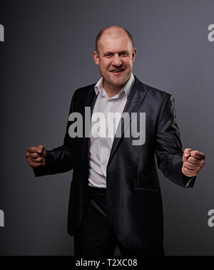 Verlassen kahlen business Mann im schwarzen Anzug, der die Hände erfolg Sieger Zeichen auf grauem Hintergrund. Closeup Portrait Stockfoto