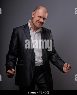 Verlassen kahlen business Mann im schwarzen Anzug, der die Hände erfolg Sieger Zeichen auf grauem Hintergrund. Closeup Portrait Stockfoto