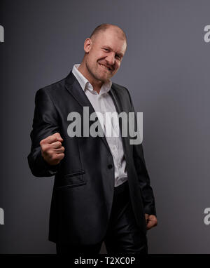 Verlassen kahlen business Mann im schwarzen Anzug, der die Hände erfolg Sieger Zeichen auf grauem Hintergrund. Closeup Portrait Stockfoto