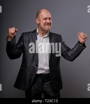 Verlassen kahlen business Mann im schwarzen Anzug, der die Hände erfolg Sieger Zeichen auf grauem Hintergrund. Closeup Portrait Stockfoto