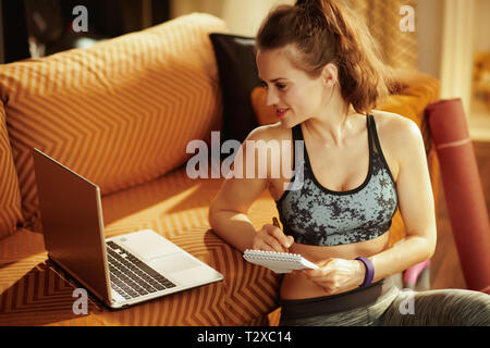 Sportlich aktive Frau in Fitness Kleidung mit Notizblock und Stift Notizen, während Sie online Streaming fitness Website in Laptop im modernen Haus. Stockfoto
