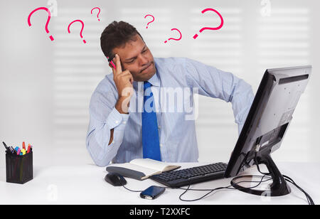 Ein Mann sitzt vor einem Bildschirm, einer Tastatur und einer Maus mit einem Sorgen, Zweifel oder Denken Ausdruck auf seinem Gesicht und Fragezeichen um seinen Er Stockfoto