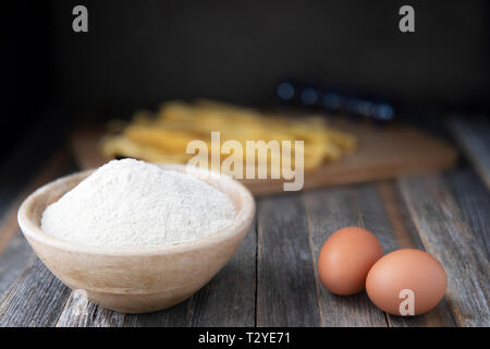 Schüssel mit Vollkornmehl und zwei Eier auf einem rustikalen Holztisch mit Pappardelle im Hintergrund. Stockfoto