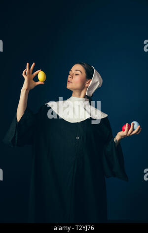 Muss perfekt sein. Mittelalterliche junge Frau als Nonne in Vintage Kleidung und weiß Mutch stehend mit Ostern Eier gefärbt Rot, Gelb und Blau Stockfoto