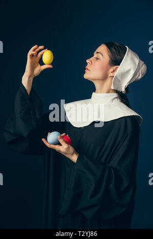 Auf der Suche nach der besten Qualität. Mittelalterliche junge Frau als Nonne in Vintage Kleidung und weiß Mutch stehend mit den farbigen Ostern rote, gelbe Eier und Stockfoto