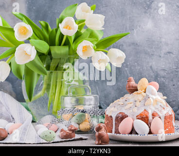 Blumenstrauß aus den weißen Tulpen in der Vase auf einem grauen Hintergrund. Shokloy Eier in verschiedenen Farben und Ostern Kuchen mit Sahne, als Symbol für die Osterferien Stockfoto