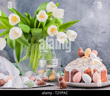 Blumenstrauß aus den weißen Tulpen in der Vase auf einem grauen Hintergrund. Shokloy Eier in verschiedenen Farben und Ostern Kuchen mit Sahne, als Symbol für die Osterferien Stockfoto