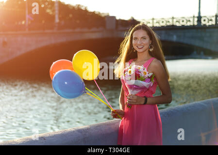 Schöne Mädchen lächelt bei Sonnenuntergang in rosa Kleid am Fluss. hübsche Frau mit Luftballons und ein Blumenstrauß. Konzept der glücklichen Stimmung schön da Stockfoto