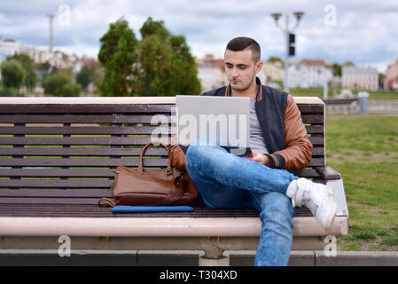 Junge männliche Freelancer arbeitet mit Laptop in der Straße auf der Bank. in der Nähe von Leder Tasche Stockfoto