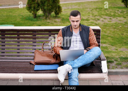 Junge männliche Freelancer arbeitet mit Laptop in der Straße auf der Bank. in der Nähe von Leder Tasche Stockfoto