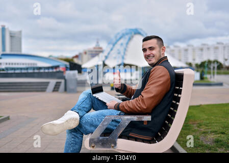 Erfolgreiche Freiberufler glücklich lächelnd. Mann in Jeans und Lederjacke auf einer Bank sitzen. jungen Mann schüttelt und zeigt Daumen hoch, wie Stockfoto