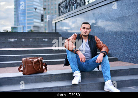 Porträt der jungen Kerl allein in Leder Jacke sitzen auf der Treppe in der Stadt. In der Nähe von Bag Stockfoto