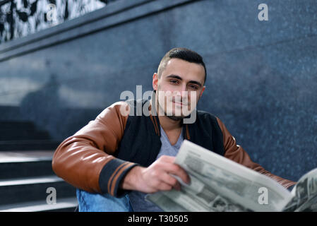 Porträt der jungen Kerl in Lederjacke lächelnd und lesen Zeitung. Mann sitzt auf der Treppe in der Stadt Stockfoto