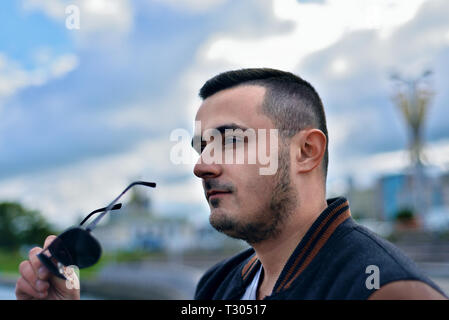 Portrait von brutalen Mann in der Stadt vor dem Hintergrund der blauen Himmel mit Wolken. jungen Kerl hält Sonnenbrille Stockfoto