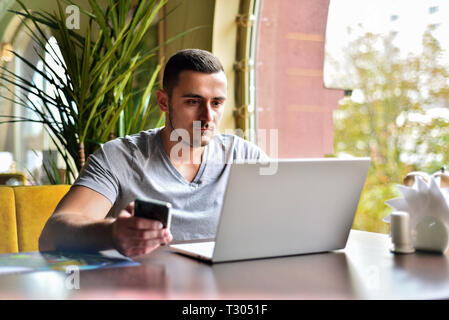 Junge Kerl ist Freelancer in Cafe hinter Laptop arbeiten. Mann hält das Telefon in der Hand und sendet Nachricht Stockfoto