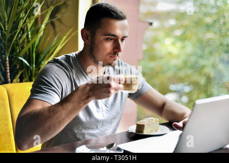 Junge Kerl ist Freelancer in Cafe hinter einem Laptop arbeiten. Mann Kaffee trinken. Es gibt Kuchen auf dem Tisch Stockfoto