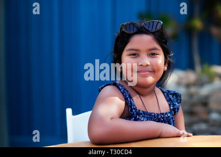 Porträt einer freundlichen kleinen Mädchen mit Schattierungen und bescheidene Kleidung beim Sitzen und Lächeln in einem Außenbereich. Stockfoto