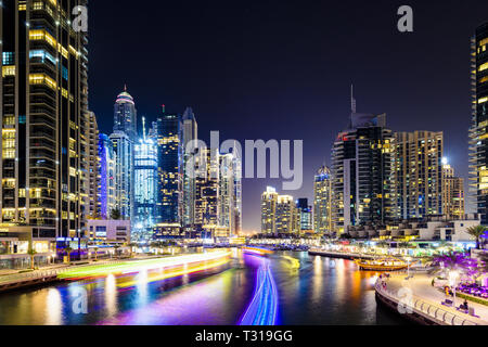 Atemberaubende Aussicht auf den Jachthafen von Dubai in der Dämmerung mit beleuchteten Wolkenkratzer im Hintergrund und einer leichten Wanderwegen durch eine Yacht segeln im Vordergrund links. Stockfoto
