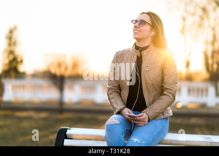 Frau hören von Musik im Freien. Glücklich lächelnde Dame sitzen auf einer Parkbank holding Handy im Sonnenuntergang. Draußen zu hören Hörbuch. Stockfoto