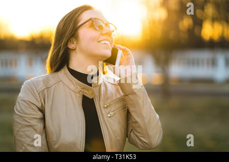 Fröhliche Frau lächelt und lacht, während sie im Freien bei Sonnenuntergang telefoniert. Junge Dame, die ein Gespräch mit einem Freund hat. Person mit Smartphone. Stockfoto