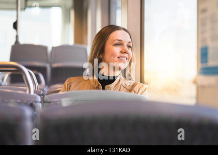 Fröhliche Dame, die in Bus, Bahn, Straßenbahn oder U-Bahn auf das Fenster blickt. Lächelnde Frau, die mit öffentlichen Verkehrsmitteln unterwegs ist, um zu arbeiten oder Urlaub zu machen. Stockfoto