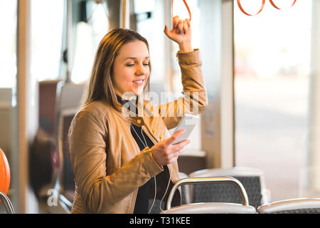 Frau, die im Zug, in der Straßenbahn oder im Bus steht, die den Griff hält und das Handy benutzt. Fröhliche weibliche Beifahrerin textet während der Fahrt mit dem Smartphone. Stockfoto