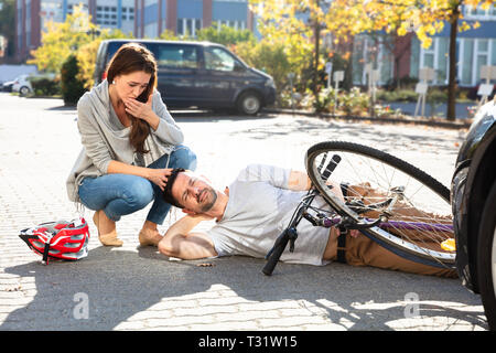 Junge Frau auf Unbewussten männlichen Radfahrer liegen an der Straße nach Unfall in der Nähe von Auto Stockfoto