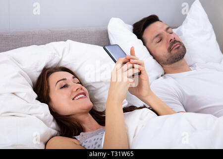 Lächelnde Frau mit Smartphone, während ihr Mann schlafend auf dem Bett im Schlafzimmer Stockfoto