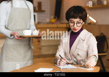 Teilweise mit Blick auf die Kellnerin holding Fach in der Nähe der trendigen Geschäftsfrau, sitzen am Tisch und Schreiben im Notebook Stockfoto