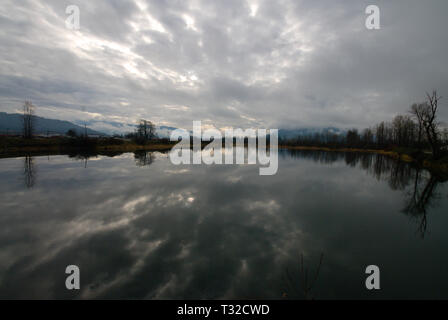 Am ruhigen Ufer des Vedder River spiegelt sich ein turbulenter grauer, bedeckter Himmel in Chilliwack, British Columbia, Kanada, wider Stockfoto