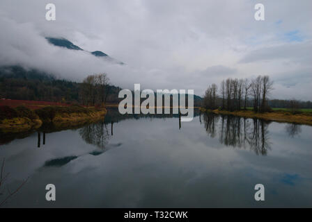 Am ruhigen Ufer des Vedder River spiegelt sich ein turbulenter grauer, bedeckter Himmel in Chilliwack, British Columbia, Kanada, wider Stockfoto