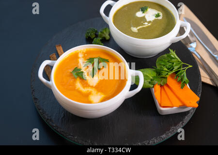 Zwei Schalen mit Kürbis und Brokkoli Suppe mit Sahne auf einem runden schwarzen Brett auf einen passenden schwarzen Hintergrund mit Servietten und Besteck serviert. Stockfoto