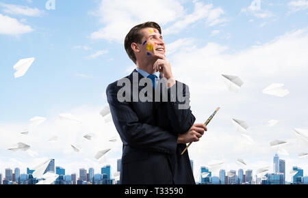 Porträt der jungen nachdenklicher Mann mit Pinsel in der Hand und Wegsehen im Stehen unter Papier fliegen Flugzeuge und gegen moderne Stadtbild Stockfoto