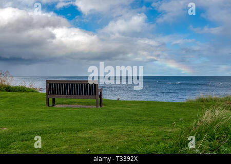 Eine Bank mit einem Regenbogen über der Nordsee Küste, im Benthall, Northumberland, England, UK gesehen Stockfoto