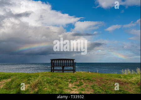 Eine Bank mit einem Regenbogen über der Nordsee Küste, im Benthall, Northumberland, England, UK gesehen Stockfoto