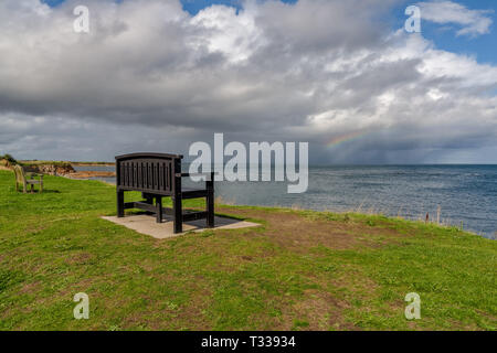 Eine Bank mit einem Regenbogen über der Nordsee Küste, im Benthall, Northumberland, England, UK gesehen Stockfoto