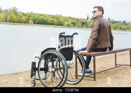 Ein junger Mann sitzt auf einer Bank am See, neben seinem Rollstuhl Stockfoto