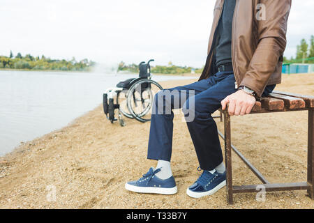 Ein junger Mann sitzt auf einer Bank am See, neben seinem Rollstuhl Stockfoto
