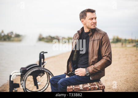 Ein junger Mann sitzt auf einer Bank am See, neben seinem Rollstuhl Stockfoto