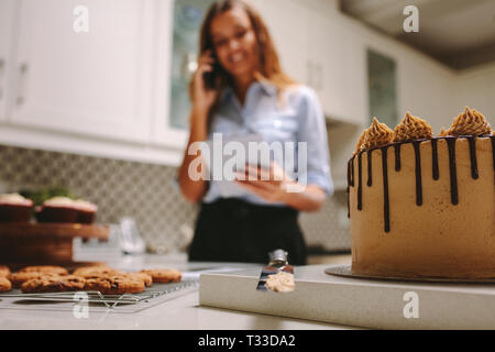 Süßwaren auf Arbeitsplatte in der Küche mit Köchin stehen im Hintergrund mit Handy und digitale Tablet. Konditor die Bestellungen am Telefon. Stockfoto