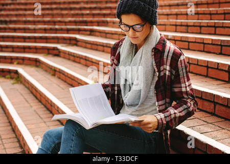 Schöne Schüler Mädchen mit Lehrbüchern auf dem College Campus. Junge kaukasier Frau in Gläsern sitzen auf der Treppe und ein Buch lesen. Stockfoto