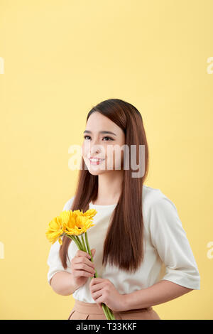 Schöne Frau im weißen Hemd mit Blumen Gerbera in den Händen auf einem gelben Hintergrund. Sie lächelt und lacht Stockfoto