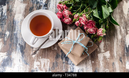 Blumenstrauß aus Rosen, Geschenke und Tee auf einem hölzernen rustikalen Hintergrund. Tag der Frauen Konzept. Ansicht von oben Stockfoto