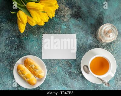 Weiße leere Karte, gelbe Tulpen, Tasse Kaffee und Kuchen auf Blau konkreten Hintergrund. Ansicht von oben. Platz kopieren Stockfoto