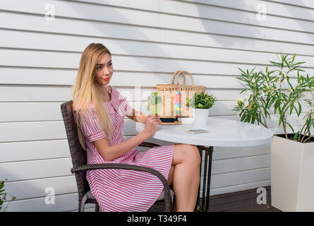 Schöne Frau in rot-weiß gestreiften Kleid. Im Sommer sitzt in einem Cafe auf der Veranda. Warten auf Freunde und Bestellung Abendessen Mittagessen Abendessen in einem Stockfoto