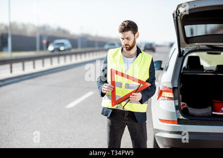 Der Mensch im Straßenverkehr Weste holding Not-Dreieck in der Nähe von Den kaputten Auto auf den Straßenrand Stockfoto