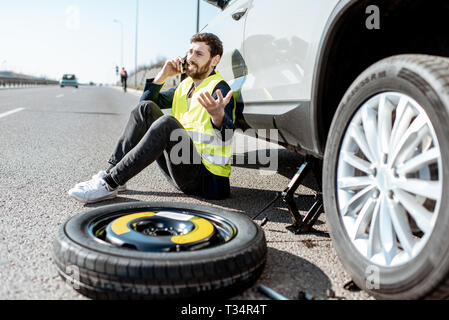 Mann Aufruf der Pannenhilfe in der Nähe des Auto sitzen mit gebrochenen Rad auf der Straße Stockfoto