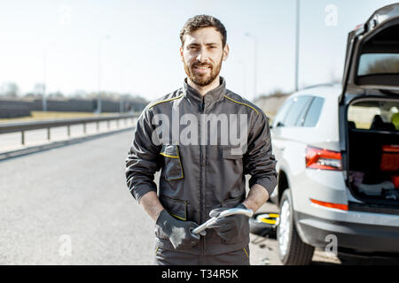 Porträt einer stattliche Straße Hilfe Arbeiter in Uniform in der Nähe von Den kaputten Auto auf der Autobahn Stockfoto