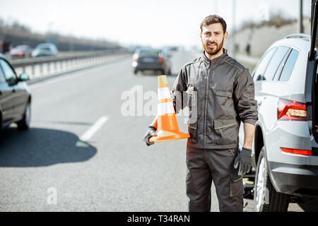 Portrait Of Happy Pannenhilfe Arbeiter in Uniform stehend mit Kegel in der Nähe des Auto auf den Straßenrand Stockfoto