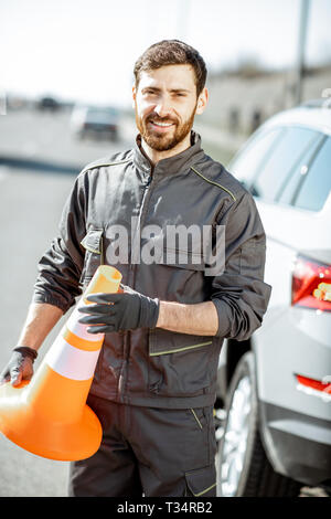 Portrait Of Happy Pannenhilfe Arbeiter in Uniform stehend mit Kegel in der Nähe des Auto auf den Straßenrand Stockfoto